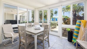 A sunlit room with a table and chairs, large windows, and a view of a pool and palm trees. - Luxury 3BR Key Largo Townhome | Ocean & Pool Views (Key Largo)