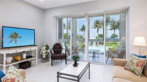 A modern living room with a television, a sofa, a coffee table, and a view of a pool and palm trees through large windows. - Luxury 3BR Key Largo Townhome | Ocean & Pool Views (Key Largo)