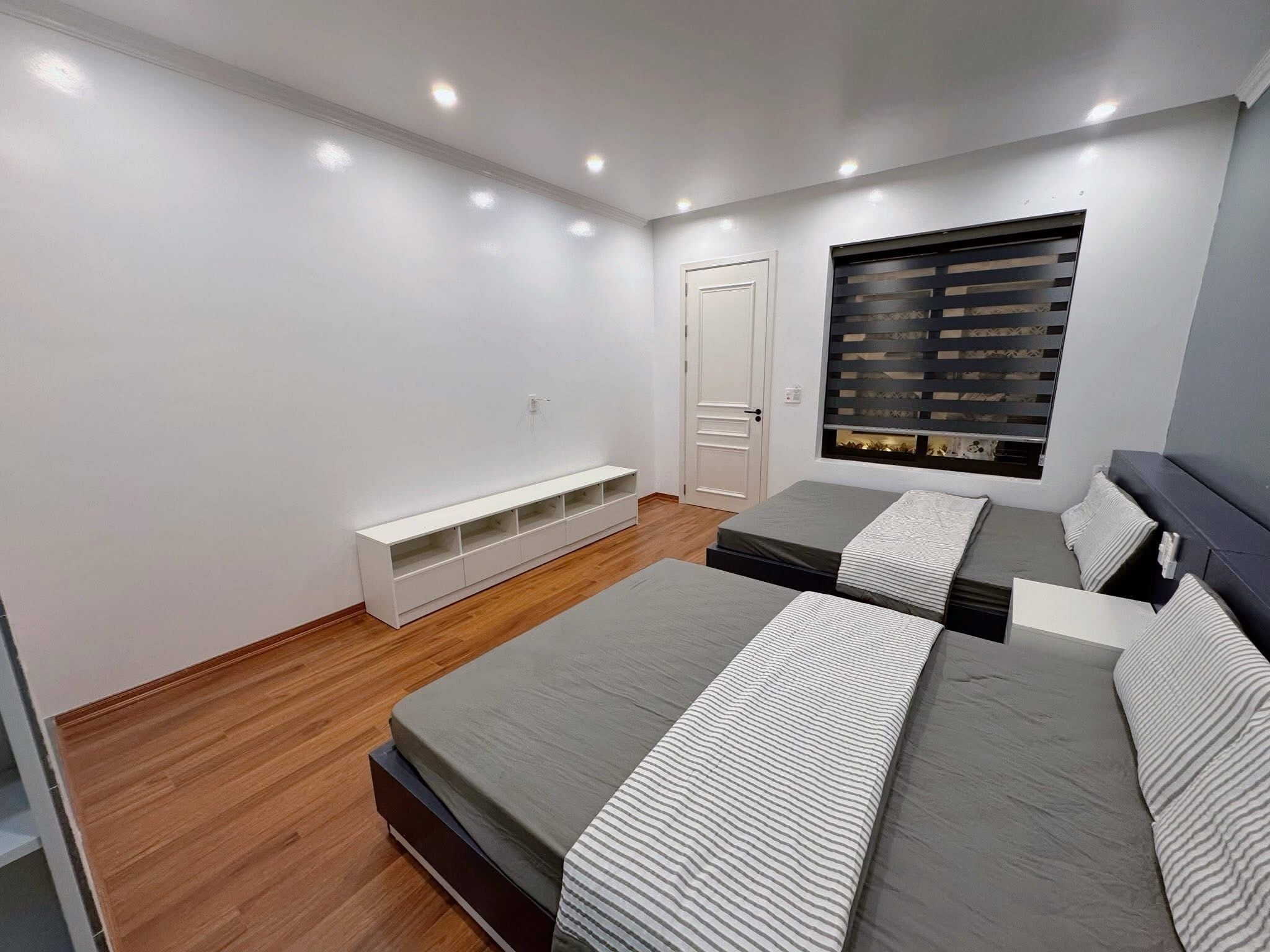 A modern bedroom with two beds, a built-in bench, a window with blinds, and recessed ceiling lights.