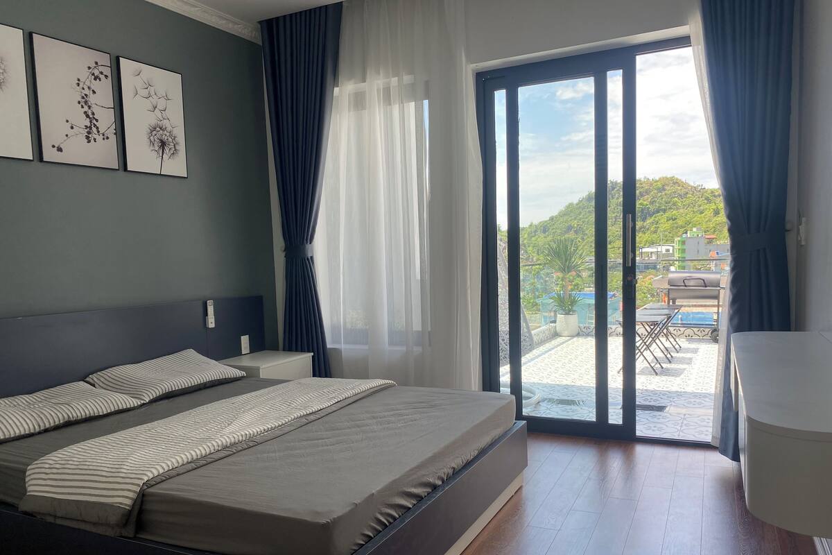 A modern bedroom with a large bed, a view of a balcony with outdoor furniture, and artwork on the wall.