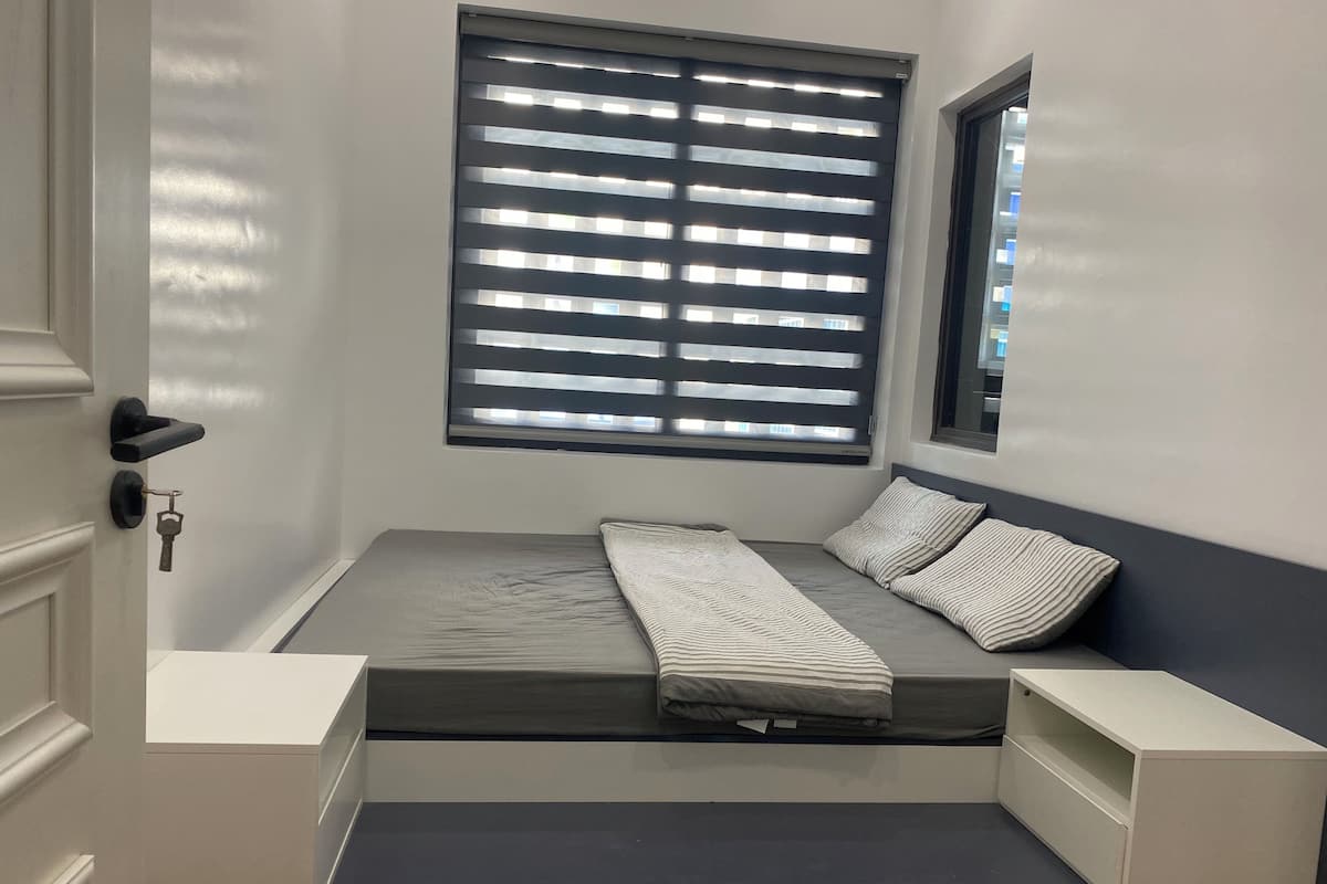 A bedroom with a bed, a window with blinds, a bedside table, and a nightstand.