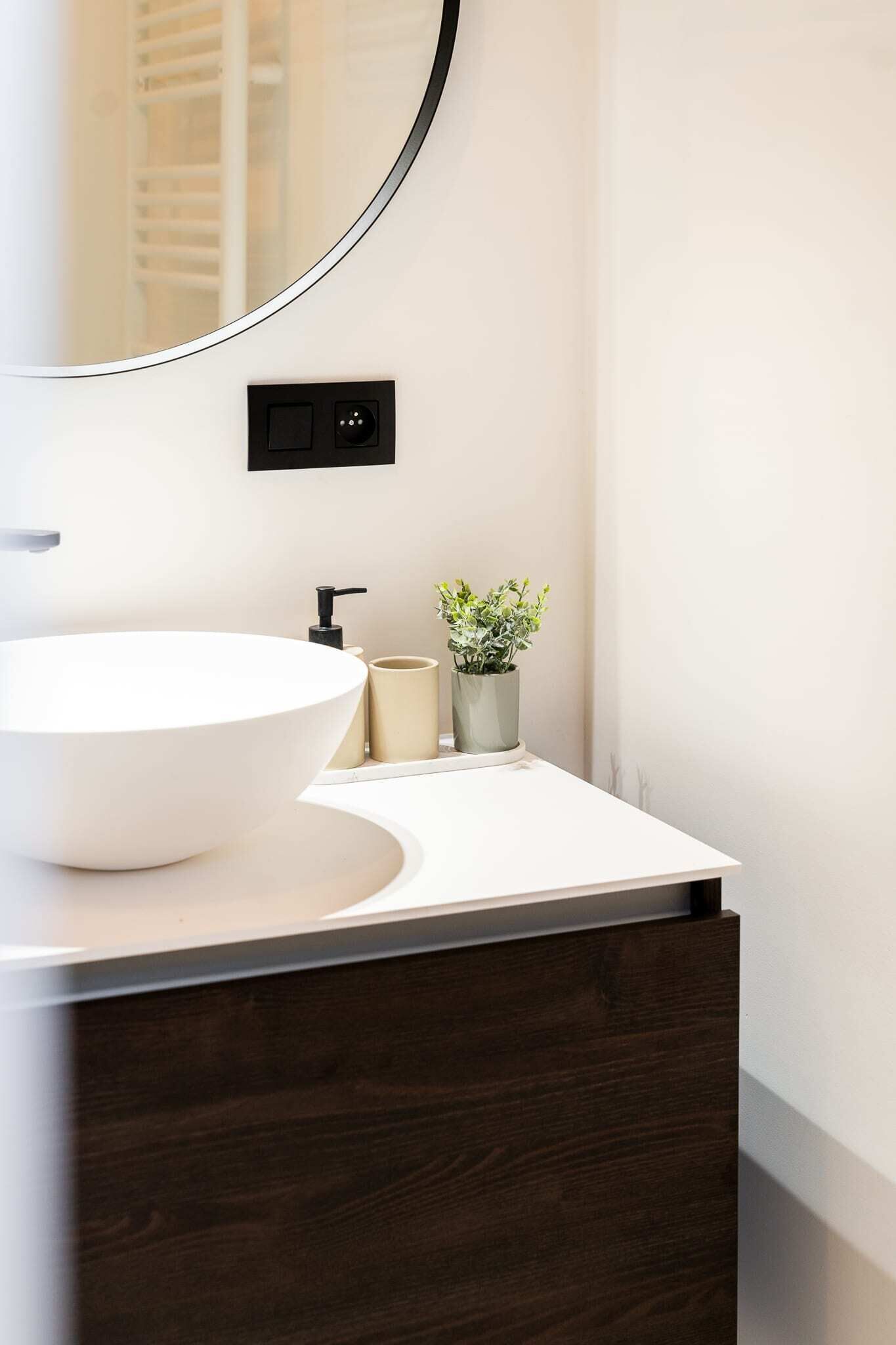 A modern bathroom with a large white vessel sink, a round mirror, a dark wood cabinet, a small potted plant, and a beige container.