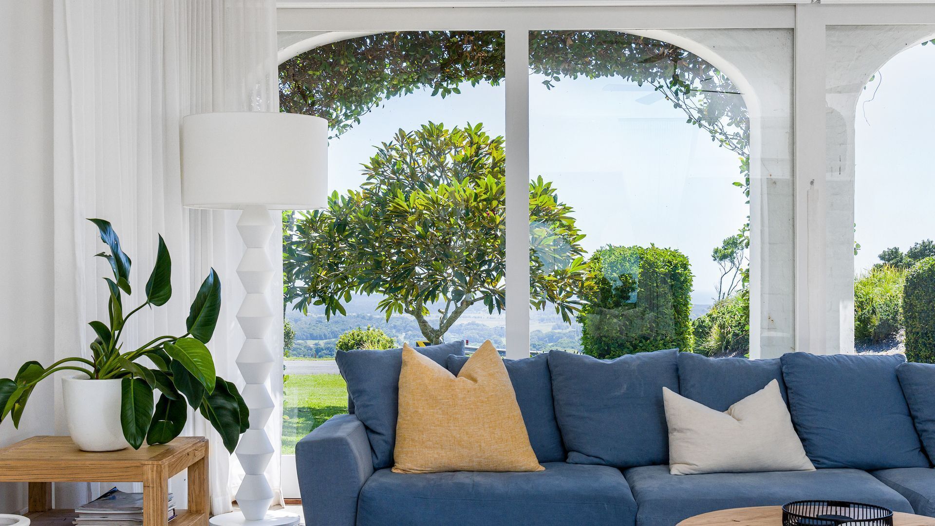 Una sala de estar moderna con un sofá azul, una lámpara blanca y vistas a un árbol y un paisaje a través de ventanas amplias.