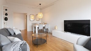Ein modernes Wohnzimmer mit grauem Sofa, einem Glastisch, einem Flachbildfernseher und einem Essbereich mit weißem Tisch und Stühlen.