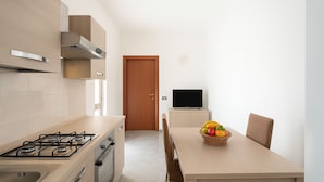 Eine moderne Küche mit Gasherd, Mikrowelle und einem Holzkasten. Ein Esstisch mit Obstkorb und einem Fernsehgerät auf einem Standbild.