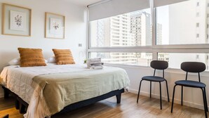 A bedroom with a bed, two pillows, a window with a view of buildings, and two chairs. - Modern Studio in Nunoa | Stylish Urban Retreat (Ñuñoa)
