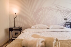 Una cama bien hecha con ropa blanca, una lámpara de noche y una pared con un diseño de hojas.