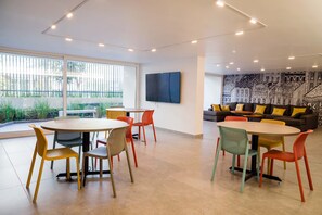 Un espace de bureau moderne avec des tables rondes et des chaises colorées, une grande fenêtre avec des stores et une fresque murale.