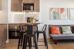 Una cocina moderna con microondas, horno y taburetes de bar. Hay una mesa de comedor con un jarrón de flores. Un sofá y obras de arte se aprecian en la sala de estar.