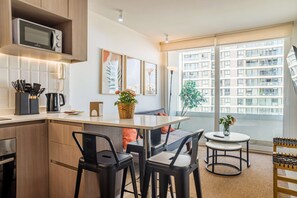 Een moderne keuken met een magnetron, koffiezetapparaat en een telefoon op het aanrecht. Er hangen kunstwerken in lijsten aan de muur, een klein tafeltje met stoelen en een uitzicht op een gebouw door grote ramen.