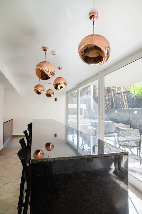 Une cuisine moderne dotée d’un grand îlot central, de luminaires suspendus en cuivre et offrant une vue sur la terrasse extérieure.