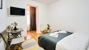Una habitación de hotel con cama, escritorio y silla, televisor montado en la pared, una mesita con planta y cortinas en la ventana.