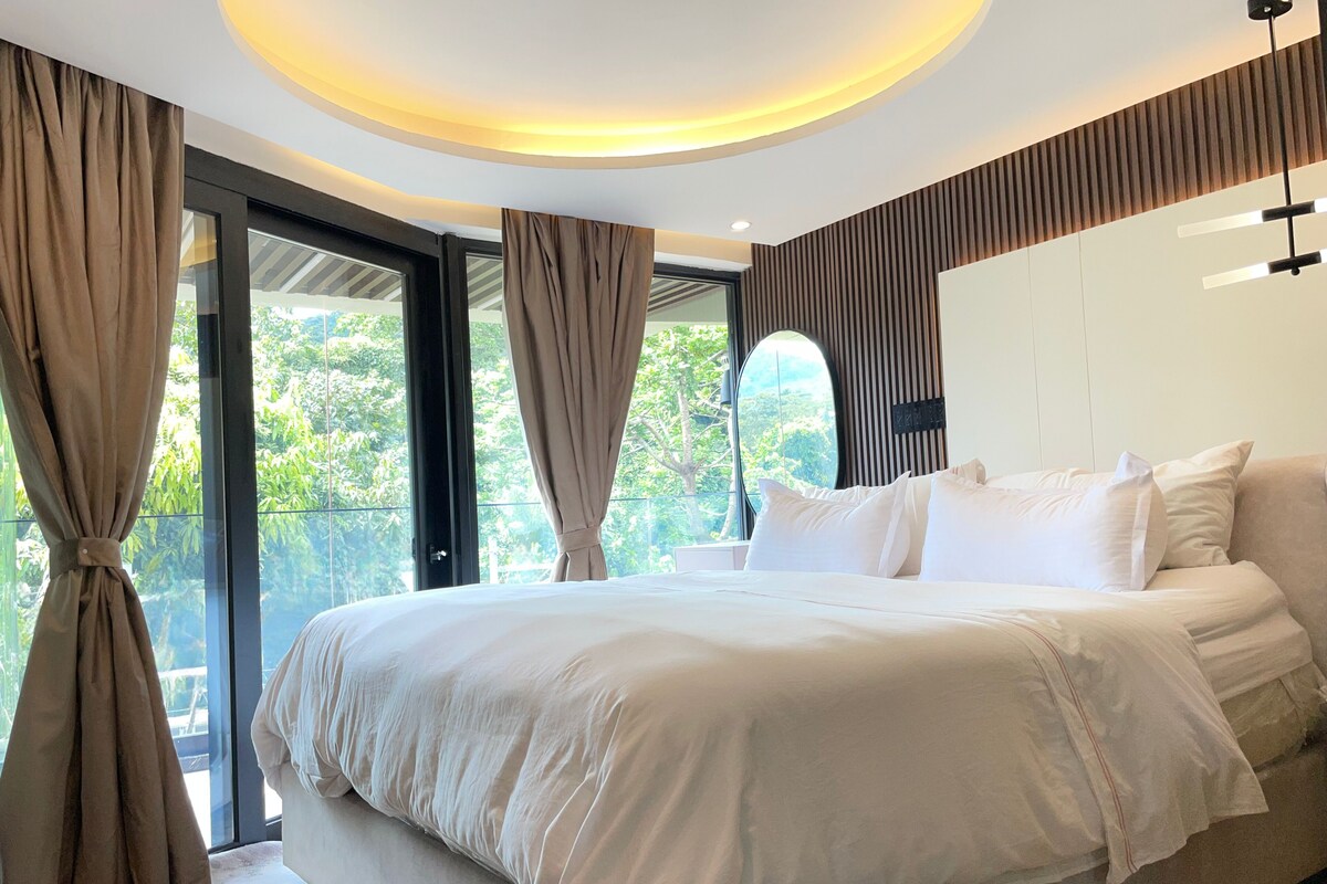A modern bedroom with a large bed, a ceiling light, and a view of greenery outside.