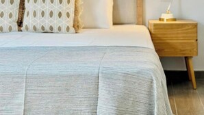 A neatly made bed with a grey textured bedspread, two patterned pillows, and a wooden headboard. A bedside table with a lamp is visible to the right.