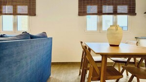 A room with a blue sofa, wooden dining table, and two windows with blinds.