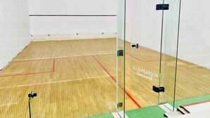 Una cancha de squash con piso de madera y paredes de vidrio, identificada como 'ELLIS PEARSON Glasswall'.