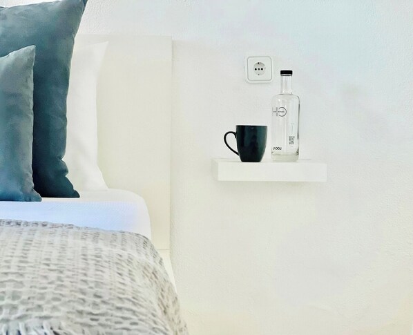 Un dormitorio ordenado con cama, una mesita de noche con una taza negra y una botella, y un enchufe en la pared.