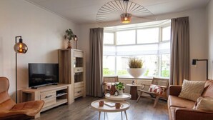 A modern living room with a flat-screen TV, wooden furniture, a round coffee table, and a large window with curtains. - Harry, Luxury property in center of NES (NES AMELAND)