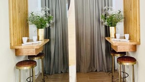 A room with a wooden partition, two stools, a small table with a vase of flowers, and a window with grey curtains.