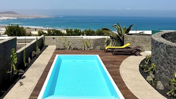 Piscina rectangular con cubierta de madera, rodeada de muros de piedra y plantas, con vistas a la costa, el océano y un paisaje urbano distante.