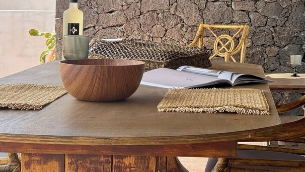 Una mesa de madera con un fondo de pared de piedra, un cuenco tejido, una botella y una silla con respaldo decorativo.