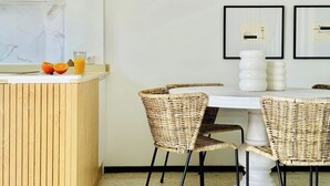 A modern kitchen with a wooden counter, wicker chairs, and a white table. There are framed pictures on the wall and a glass of orange juice on the counter. - Canaryislandshost I Marina (Barranco de la Verga)