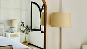 A hotel room with a large ornate mirror, a bedside lamp, a bed with a white comforter, a small potted plant, and a white nightstand with a lamp and decorative items. - Canaryislandshost I Marina (Barranco de la Verga)