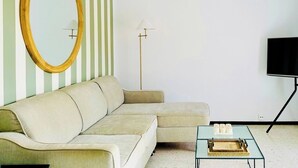 A modern living room with a striped wall, a large round mirror, a beige sofa, a floor lamp, a TV, and a small table with decorative items. - Canaryislandshost I Marina (Barranco de la Verga)