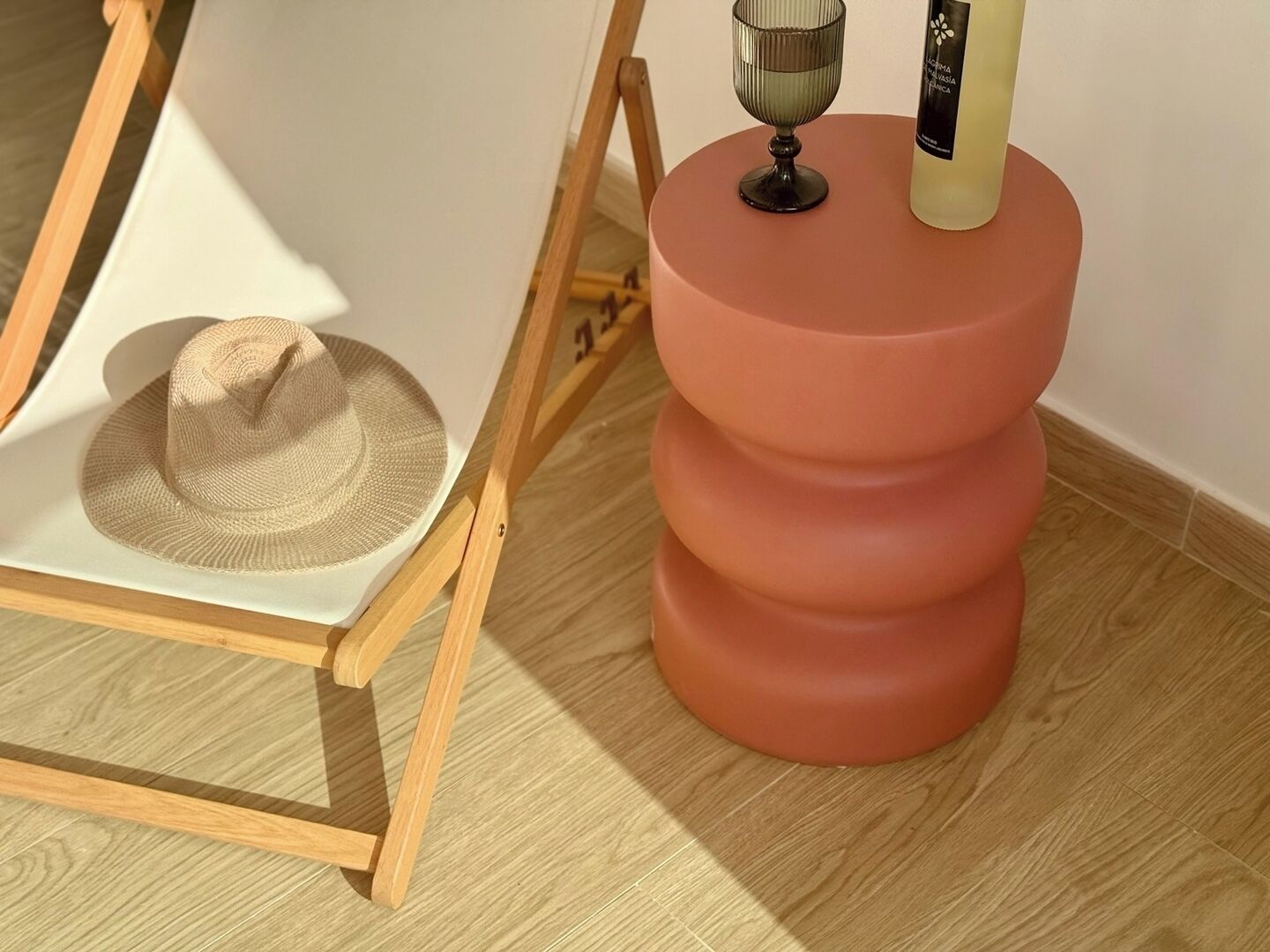 Une chaise en bois surmontée d’un chapeau de paille, un verre posé sur une petite table et un objet rose, grand et composé de plusieurs niveaux.