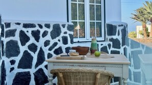Un bâtiment blanc avec un mur en pierre et une fenêtre. On y voit une table sur laquelle se trouvent un bol, une bouteille et une pomme.