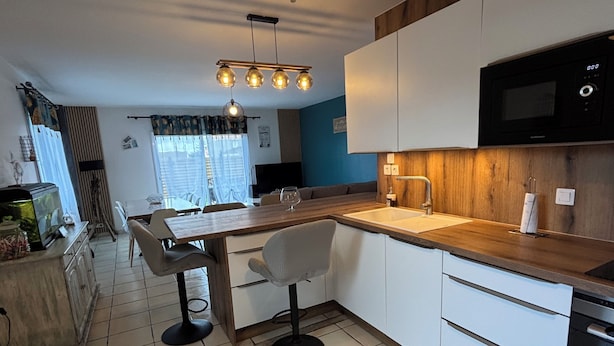 Una cocina moderna con encimera de madera, armarios blancos y microondas negro. Hay un comedor con mesa y sillas, y un amplio ventanal con cortinas.