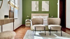 Ein modernes Schlafzimmer mit grüner Akzentwand, zwei abstrakten Gemälden, einem Bett mit passenden Kissen, zwei Polsterstühlen, einem Nachttisch mit Lampe und einem Fenster mit Jalousien.