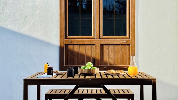 Ein Holztisch mit Frühstücksgegenständen und einer Orangensaftflasche vor einer Holztür mit Fensterfeldern.