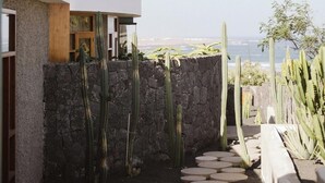 A modern outdoor patio with a stone wall, cacti, and a view of the ocean. - Canaryislandshost I Natural II (Urbanización Famara)