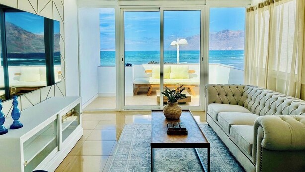 Een moderne woonkamer met een grote televisie, een bank en een salontafel. Er is een schuifdeur naar een balkon met uitzicht op de oceaan.