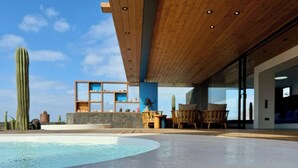 Un área de piscina moderna con techo de madera, una zona de estar con sillas y un cactus al fondo.