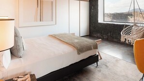 Une chambre avec un lit, un fauteuil suspendu, un mur en pierre et une fenêtre donnant sur un beau paysage.
