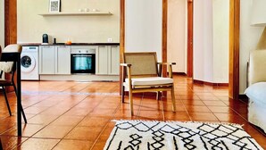 Una habitación con sofá, sillón, alfombra, lavadora y una zona de cocina con armarios y estufa.