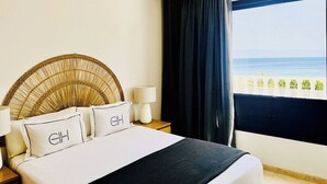 Habitación de hotel con cama, mesita de noche con lámpara, ventana grande con vista a la playa y cabecero decorativo.