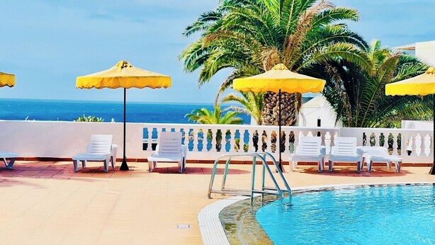 Zona junto a la piscina con sillas de descanso blancas, sombrillas amarillas y vista al océano.