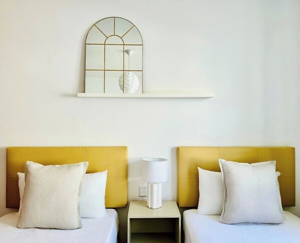 Habitación de hotel con dos camas, cada una con cabecero amarillo y ropa de cama blanca. Entre las camas, una mesita de noche blanca con una lámpara. Encima de las camas, hay un estante blanco con un espejo decorativo.