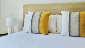 Un lit bien fait, agrémenté d’oreillers blancs et moutarde, une lampe de chevet blanche et un mur sobre orné d’une œuvre d’art à la texture intéressante.