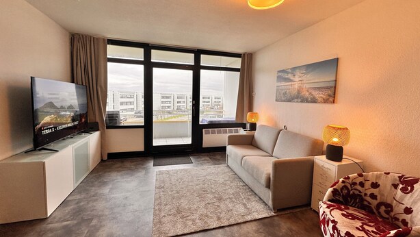 Een moderne woonkamer met een televisie, bank en fauteuil, grote ramen en een plafondlamp.