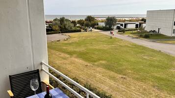 A balcony with a table set for two, overlooking a grassy area with a view of the sea and a building in the distance.