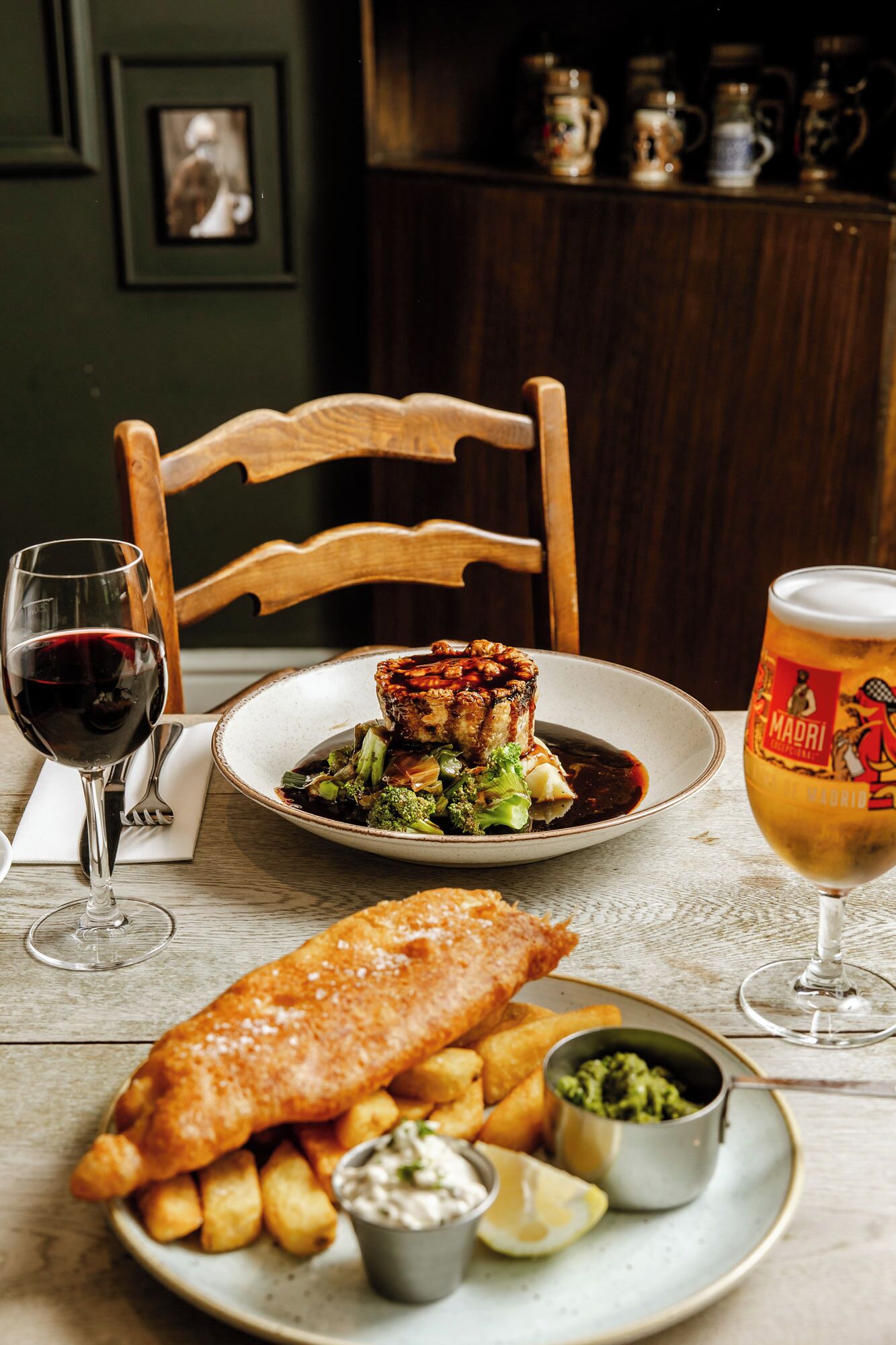 A dining table with a plate of fish and chips, a glass of beer, and a glass of red wine.