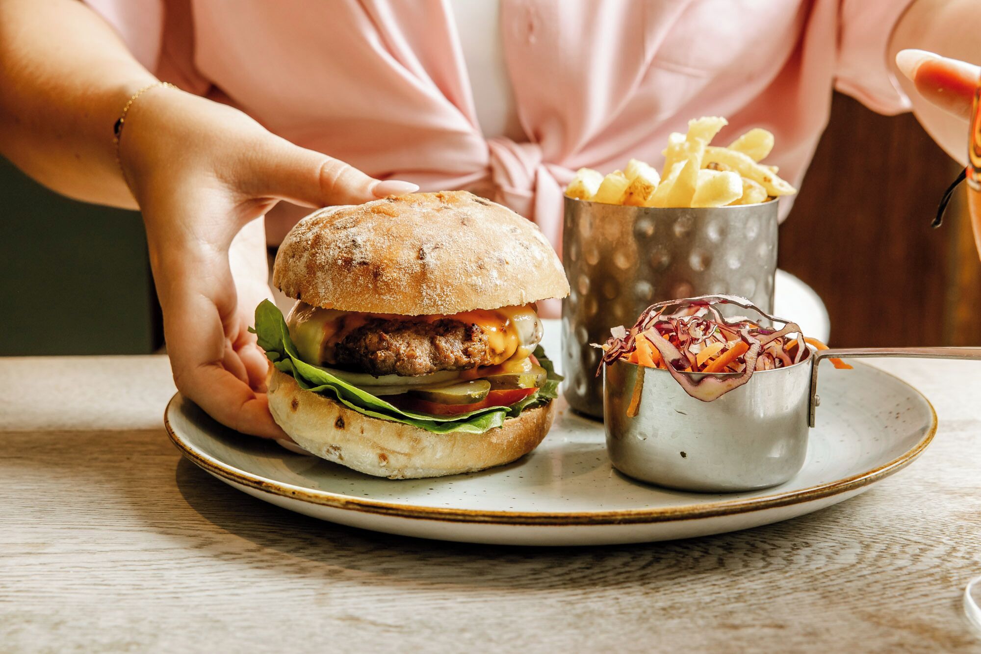 A person holding a burger with fries and coleslaw on a plate.