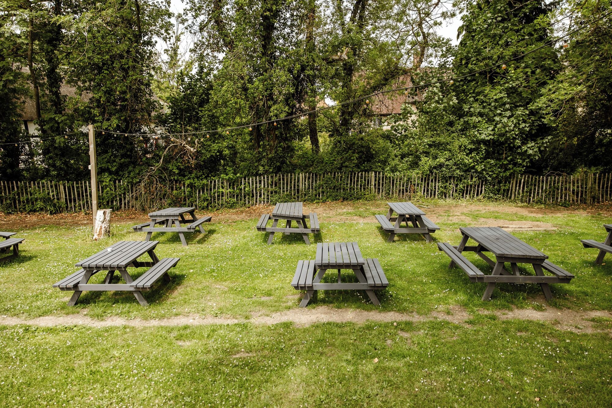 Ett utomhusområde med picknickbord placerade i en cirkel, omgivet av ett trästaket och tät grönska.