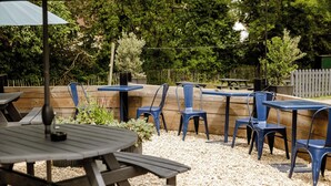 An outdoor patio area with wooden tables and blue plastic chairs, surrounded by a wooden fence and greenery. - The Bell Inn (Ladbroke)