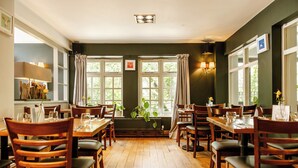 A restaurant with wooden tables and chairs, large windows with curtains, and a green wall. - The Bell Inn (Ladbroke)