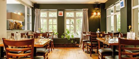 Un restaurante con mesas y sillas de madera, amplias ventanas con cortinas y una pared verde.